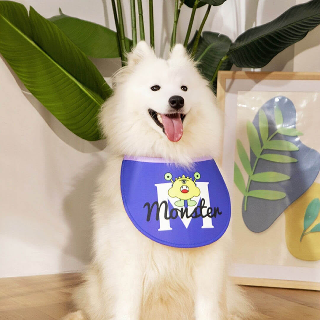 A big white samo dog wears a blue adjustable dog drool bib with little monster printing sitting on the floor in front of a big green plant