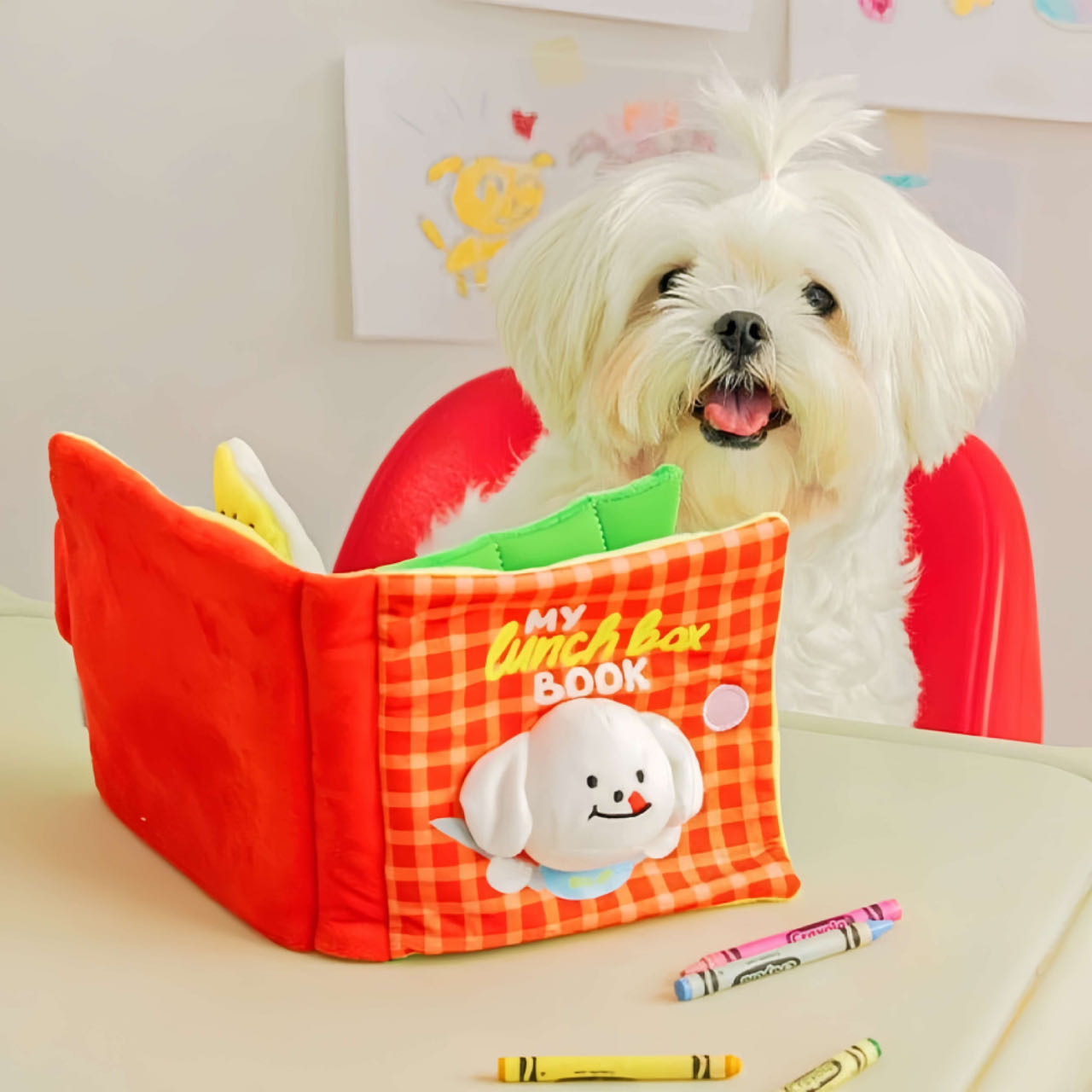 A Red Colour Dog Snuffle Toy With Book Style Standing  On The Table In Front Of A White Puppy Dog 